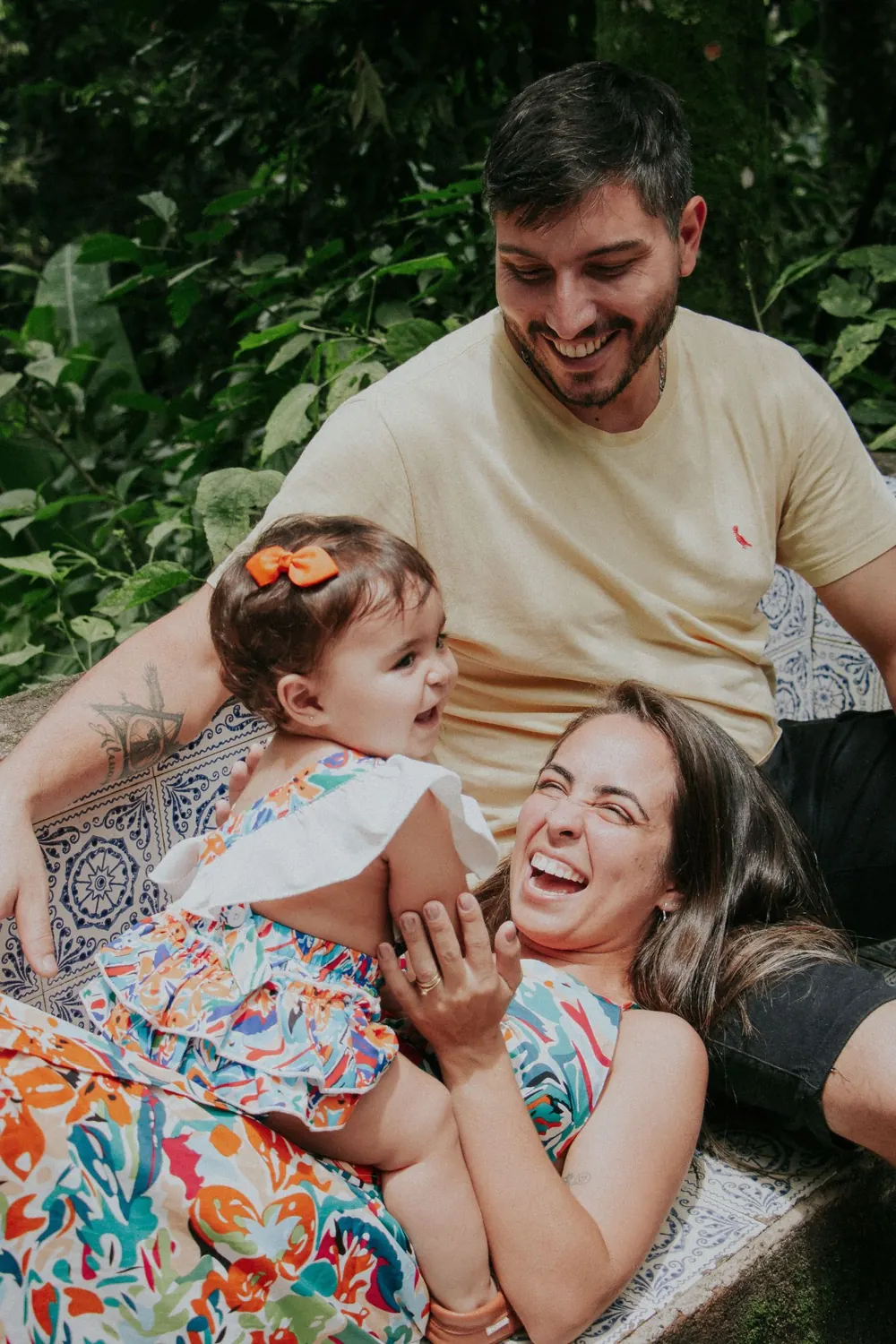 Casal rindo com a filha pequena no colo, sentados em um banco de azulejos entre plantas